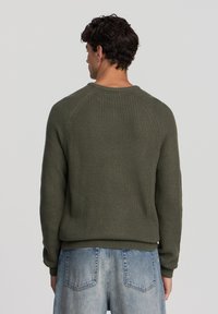 Maglione lavorato a maglia verde oliva con texture a coste, scollatura rotonda e maniche raglan. Polsini e orlo a coste. Indossato con pantaloni corti di denim blu sbiadito.