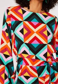 Mehrfarbige Bluse mit einem geometrischen Muster aus Rauten und Dreiecken. Der Stoff ist glatt, mit langen Ärmeln und einer Taille zum Binden.