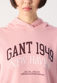 Mlada osoba koja nosi svijetloružičasti duksericu s natpisom "GANT 1949 New Haven" i "American Sportswear" na prednjoj strani.