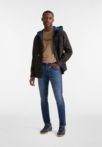 Chaqueta de cuadros oscuros con capucha azul, camiseta color beig con un gráfico de un perro, jeans azules y zapatillas navy con suelas marrones.