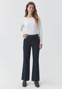 Jeanși evazați din denim albastru, cu talie înaltă, prevăzuți cu închidere prin buton și buzunare frontale, asortați cu un top alb cu mâneci lungi.