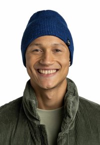 Blue knitted beanie with ribbed texture and small logo tag; worn with a green corduroy jacket, featuring a high collar and a plain shirt underneath.
