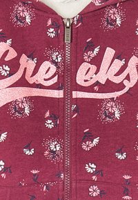 Burgunderfarbene Zip-Hoodie mit floralen Mustern und einem glitzernden pinken "Creeks"-Logo. Er verfügt über eine glatte Textur und einen mittigen Reißverschluss.