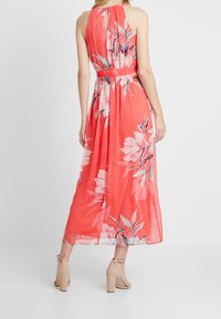 Robe maxi florale corail avec encolure halter et taille cintrée. Tissu transparent avec de grands motifs floraux roses et verts, dotée d'une découpe en forme de keyhole au dos.