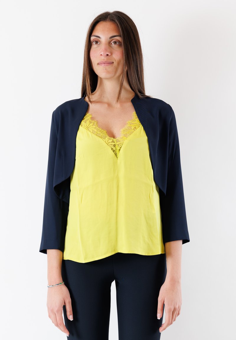 Giacca cropped blu navy sopra una canotta gialla brillante con bordo in pizzo. La canotta ha un collo a V e una texture liscia. I leggings neri completano il look.