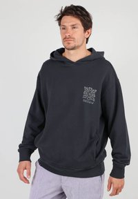 Sweat-shirt à capuche noir en tissu doux, avec une poche avant et un design géométrique sur la poitrine gauche. Coupe décontractée avec des poignets en bord-côte.