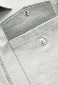 Light gray fabric featuring a textured surface, with a snap closure and a front pocket. The design includes a zipper along one side.