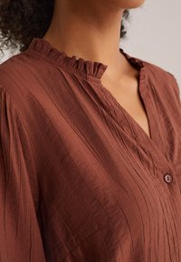 Blouse marron texturée avec un col froncé, fermeture à boutons et rayures verticales. Le tissu semble léger et légèrement froissé.