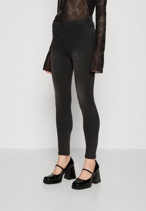 Persoon draagt hooggetailleerde, vervaagde zwarte skinny jeans, doorschijnende zwarte top met patronen, en zwarte lakleren Mary Jane blokhakken.