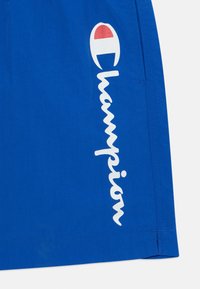 Modré športové šortky vyrobené z ľahkého materiálu, na boku s vertikálnym bielym logom "Champion" a malou nášivkou s logom.