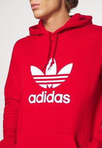 Červená mikina s kapucí vyrobená z bavlny, s velkým bílým logem Adidas a designem tří paralelních pruhů na hrudi. Šňůrky na kapuci.