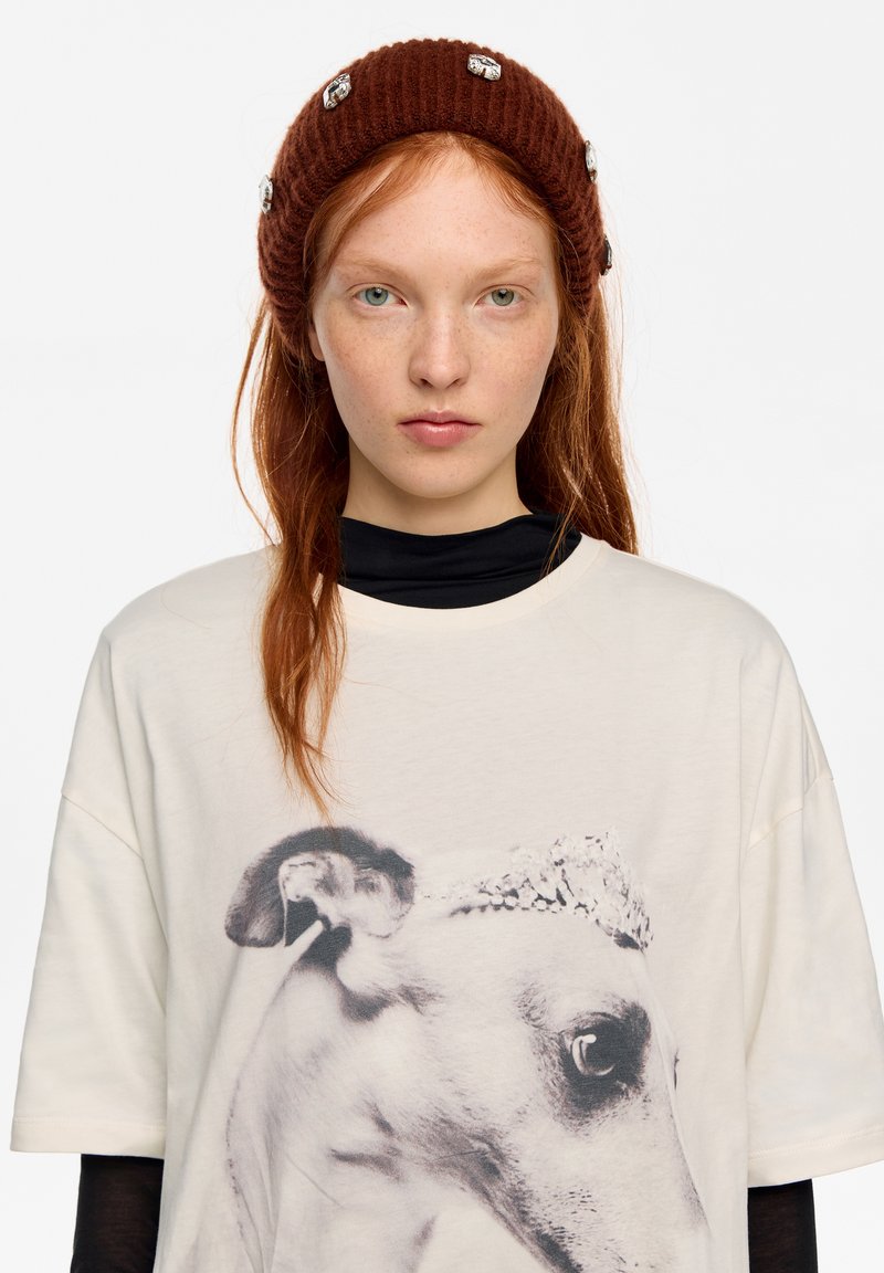 Gorro de punto marrón acanalado con detalles plateados, usado sobre una camiseta oversize color crema que presenta un gráfico de perro en blanco y negro. Cuello alto negro en capas debajo.