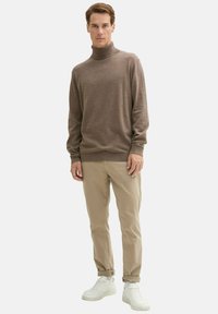 Brązowy sweter z golfem z miękkiego materiału, w zestawie z beżowymi chinosami i białymi trampkami. Sweter ma dopasowany dekolt i długie rękawy.