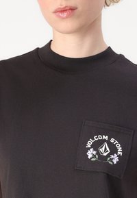 Black short-sleeve t-shirt with a crew neck, featuring a front pocket adorned with "Volcom Stone" and floral graphics in white and purple.