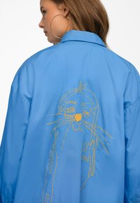 Blue oversized shirt featuring embroidered yellow cat design on the back. Made from lightweight fabric with a collar and smooth texture.