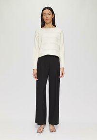 Weißer strukturierter Pullover mit Wellenmuster, kombiniert mit schwarzen weit geschnittenen Hosen und silbernen offenen Sandalen. Modell steht vor einem neutralen Hintergrund.