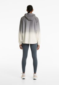 Giacca leggera con cappuccio in gradazione di grigio a bianco, con polsini elastici e vestibilità comoda, abbinata a leggings blu e scarpe da ginnastica.