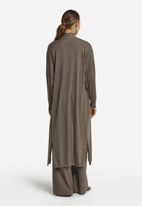 Lång, öppen cardigan i brunt, tillverkad av mjukt material, med sidofickor och långa ärmar, bärs över vida bruna byxor.