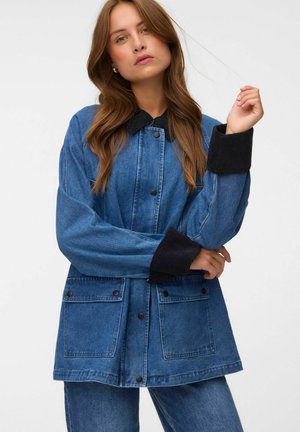 Femme aux longs cheveux bruns portant une veste en jean bleue avec un col et des poignets noirs, debout devant un fond uni.