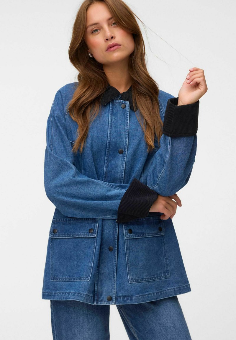 Femme aux longs cheveux bruns portant une veste en jean bleue avec un col et des poignets noirs, debout devant un fond uni.