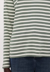 Zeleno-bíle pruhovaná košile s dlouhým rukávem, vyrobená z měkké bavlny. Vybavená zaobleným lemem a úzkými manžetami, spárovaná s tmavě modrými džínami.