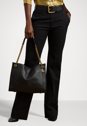 Borsa tote in pelle nera con manici a catena dorati, dotata di un'etichetta con il logo. Texture liscia con una forma strutturata, tenuta da una persona che indossa pantaloni neri.