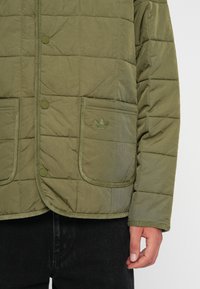 Alyvuogių žalios spalvos pūkinė striukė su siuvinėjimu, spaudėmis mygtukais, dviem priekinėmis kišenėmis ir toniniu Adidas Trefoil logotipu ant krūtinės.