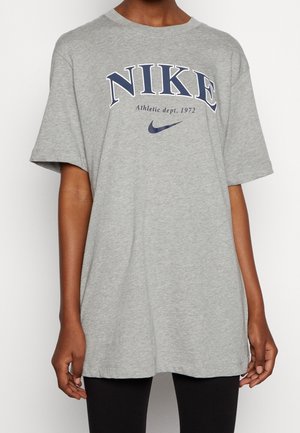 Osoba nosząca szarą koszulkę Nike z tekstem i logo "NIKE Athletic dept. 1972", w czarnych spodniach, stojąca na białym tle.