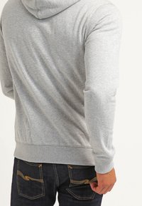 Felpa grigia con cappuccio, realizzata in una morbida miscela di cotone, con orlo e polsini a costine. Sullo sfondo si vedono jeans di denim con cuciture gialle.