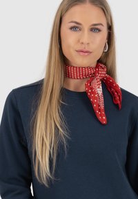 Rode stippen sjaal om de nek geknoopt, gemaakt van een lichte, gestructureerde stof, contrasterend met een donkerblauwe sweatshirt.