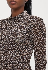 Robe à imprimé léopard, en tissu transparent, col haut, manches longues avec épaules bouffantes, aux tons terreux de marron, noir et crème.