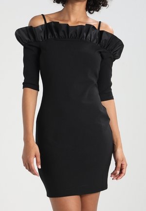 Femme portant une robe noire ajustée avec épaules dénudées, un col à volants et des manches courtes, sur un fond clair uni.