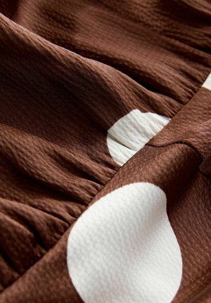 Hnědá texturovaná tkanina s velkými bílými puntíky, s jemnými shrnutými záhyby a hladkými, plynulými záclonami.