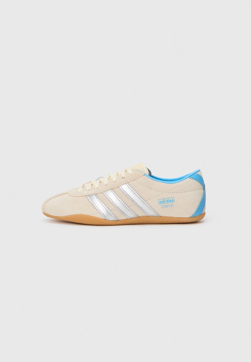 Beige semsket sneaker med sølvstriper, blå detaljer på hælen og tungen, og en gummisåle. "adidas TOKYO" logo på siden.