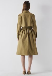 Mujer vistiendo un vestido midi verde olivo con mangas largas, cintura fruncida y una capa corta, de pie descalza frente a un fondo blanco liso.