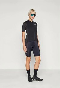 Μαύρη ποδηλατική φανέλα με κοντά μανίκια, φερμουάρ μπροστά, σε συνδυασμό με μαύρα σορτς. Τα αξεσουάρ περιλαμβάνουν γυαλιά ηλίου με μπλε τόνους και μαύρα ποδηλατικά παπούτσια.