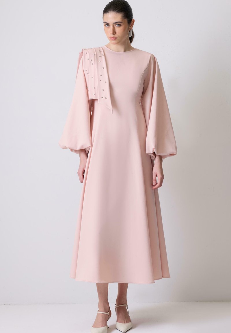 Γυναίκα με μαλλιά δεμένα πίσω φοράει ένα μακρύ ανοιχτό ροζ φόρεμα με μπαλόν μανίκια και ένα διακοσμημένο κασκόλ που κρέμεται πάνω σε έναν ώμο, στέκεται μπροστά σε λευκό τοίχο.