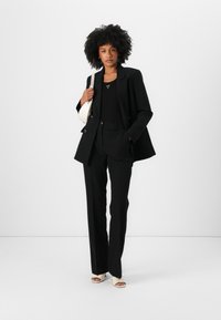 Svart skreddersydd blazer over en tettsittende svart topp, kombinert med høytlivte svarte bukser og beige høye hæler, mens hun holder en hvit veske.