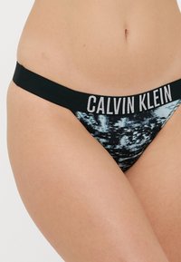 Calvin Klein juosmens bikini kelnaitės juodos spalvos su mėlynu abstrakčiu raštu, pasižyminčios minkštu juosmeniu, ant kurio užrašyta prekės ženklas baltais rašmenimis.