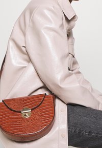 Donna che indossa una giacca beige lucida e jeans grigio scuro, con una borsa a tracolla marrone in pelle effetto coccodrillo con chiusura dorata.