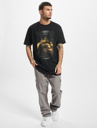 T-shirt graphique noir avec un grand design de couronne, pantalon cargo gris et baskets blanches. Le modèle se tient devant un fond neutre, mettant en valeur les détails de la tenue.