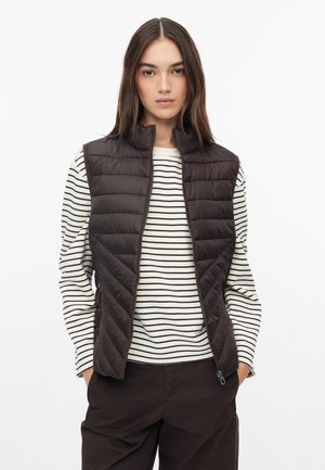Junge Frau trägt eine braune gesteppte Weste über einem weißen und schwarzen gestreiften Langarmshirt mit braunen Hosen, schaut nach vorne und hat die Hände in den Taschen.