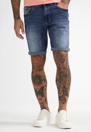 Denim shorts in mediumblauw met omgeslagen zoom; voorzien van vijf zakken. Combinatie met witte sneakers; benen versierd met verschillende tatoeages.