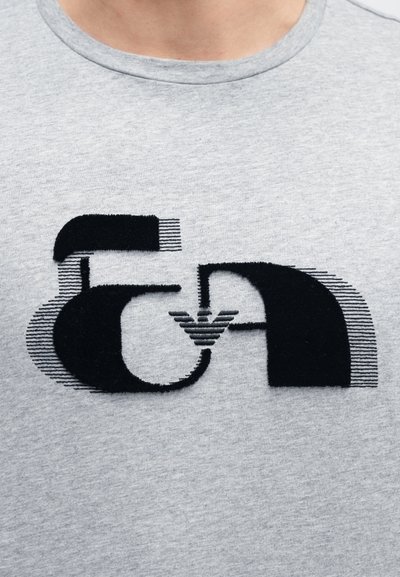 Sivá bavlnená tričko s veľkým čiernym grafickým motívom obsahujúcim abstraktné tvary a lineárne vzory, zdôrazňujúcim textúru a dizajn.