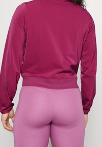 Persona con una camiseta de manga larga magenta y leggings morados claros, vista desde atrás contra un fondo sencillo.