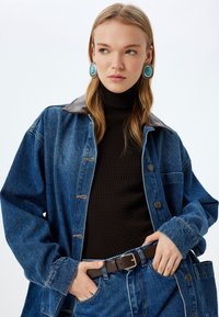 Jeansjacka med krage i brunt läder, med knäppning och fickor. Svart ribbad polotröja, brunt bälte och stora turkosa örhängen.