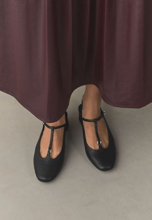Piedi con ballerine nere con cinturino a T, in piedi su pavimento grigio, parzialmente coperti da una lunga gonna marrone.