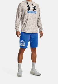 Mężczyzna ubrany w szary kaptur Under Armour, niebieskie spodenki Under Armour, białe skarpetki i białe buty sportowe, stojący na białym tle.