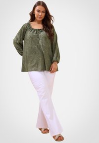 Olivgrüne Bluse mit einem strukturierten Zebra-Muster und quadratischem Ausschnitt, kombiniert mit weißen Schlaghosen und Sandalen mit metallischen Akzenten.