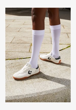 Baskets blanches de sport avec une surface texturée, des accents noirs et une semelle en caoutchouc marron, accompagnées de chaussettes blanches montantes.